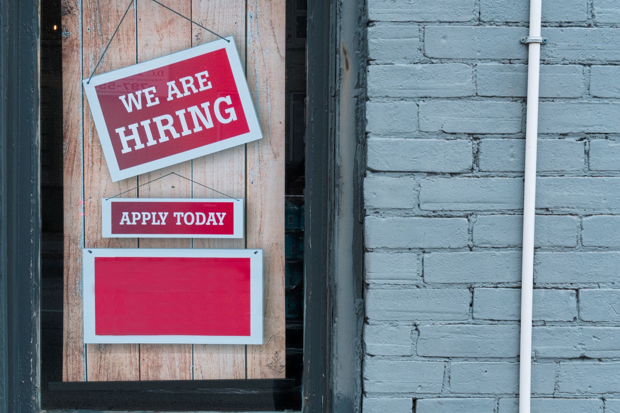 Two red signs outside a brick building, saying "We Are Hiring" and "Apply Today"
