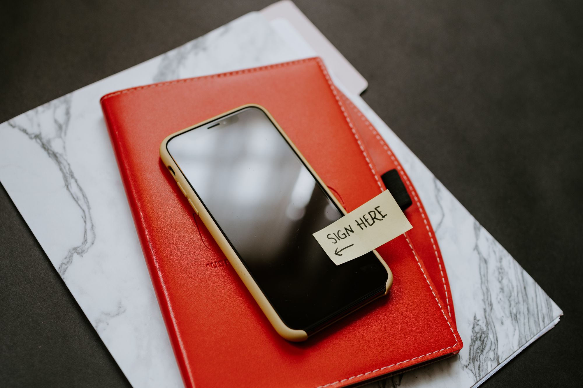Mobile phone with a "Sign Here" sticky note on it, resting on a red notebook cover and a marbleized file folder